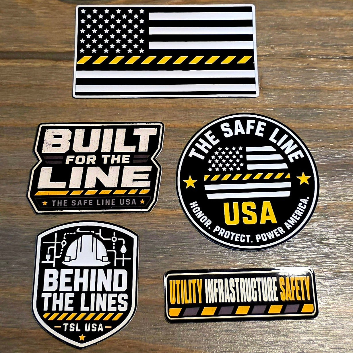 Five utility infrastructure safety stickers with black, white, and yellow designs are arranged on a rustic wooden surface. The stickers feature American flag motifs, industrial themes, and text including 'BUILT FOR THE LINE', 'BEHIND THE LINES', 'THE SAFE LINE', and 'UTILITY INFRASTRUCTURE SAFETY'.