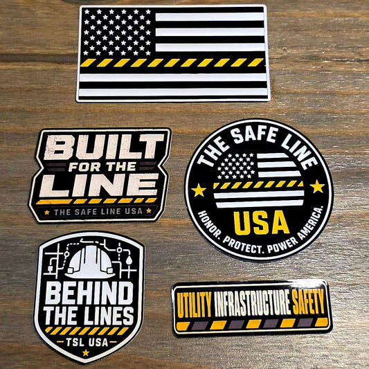 Five utility infrastructure safety stickers with black, white, and yellow designs are arranged on a rustic wooden surface. The stickers feature American flag motifs, industrial themes, and text including 'BUILT FOR THE LINE', 'BEHIND THE LINES', 'THE SAFE LINE', and 'UTILITY INFRASTRUCTURE SAFETY'.