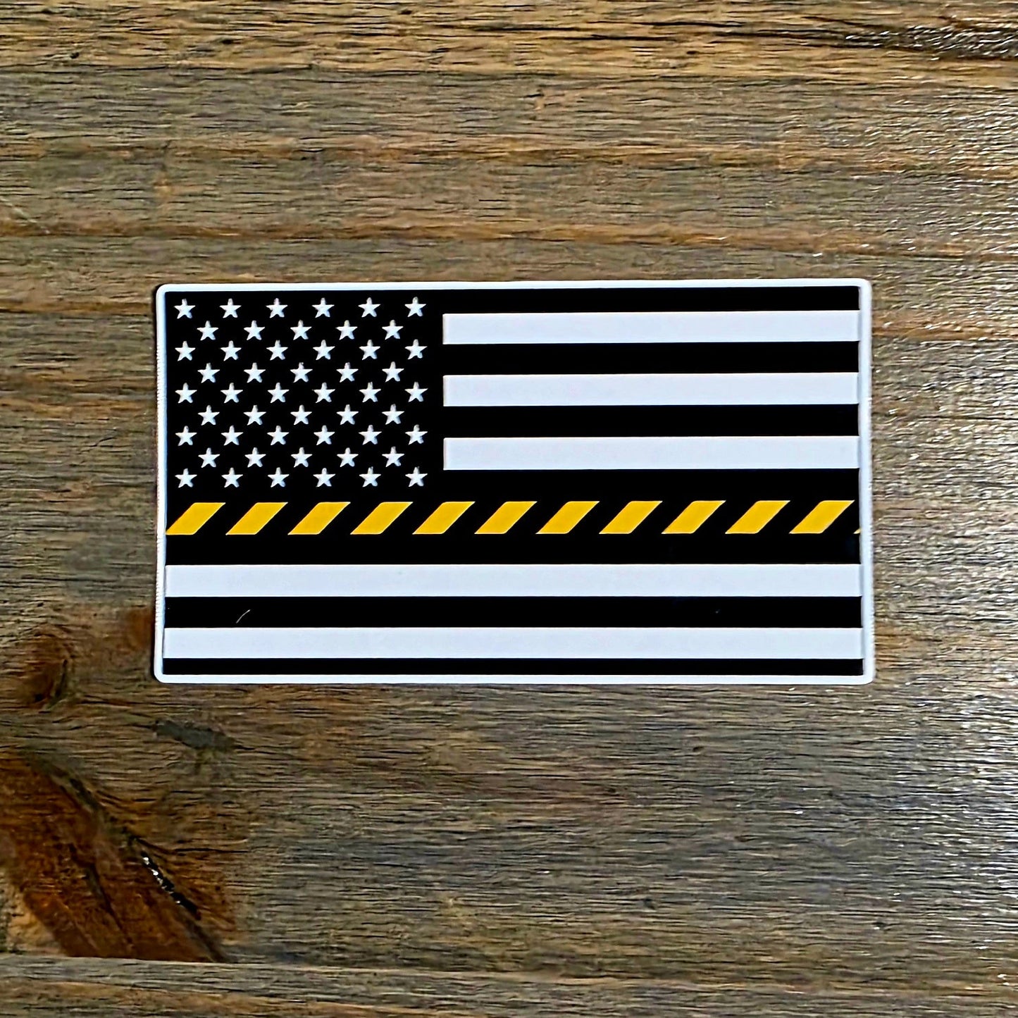 A rectangular sticker featuring a black and white American flag with a single yellow stripe across the center, placed on a dark wood surface.