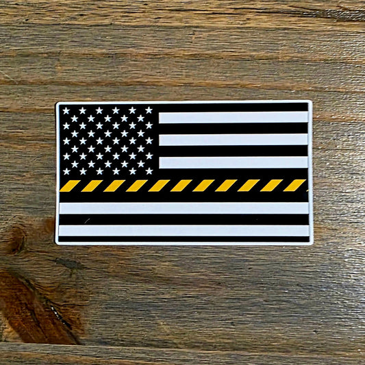 A rectangular sticker featuring a black and white American flag with a single yellow stripe across the center, placed on a dark wood surface.